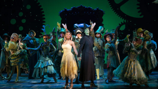 Two lead performers stand hand in hand center stage, surrounded by a large ensemble in colorful, Victorian-steampunk costumes during a theater finale.