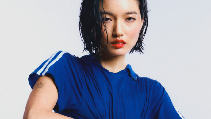 Portrait of a woman with wet black hair, wearing a blue shirt, arms crossed against a pale gradient background.