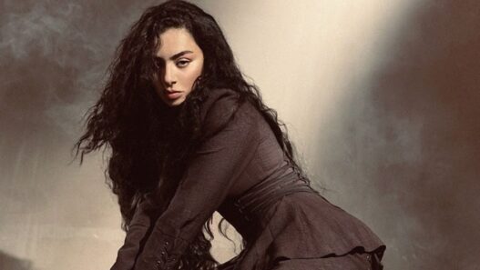 Woman with long dark curly hair in a brown coat, leaning forward against a muted studio backdrop.