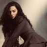 Woman with long dark curly hair in a brown coat, leaning forward against a muted studio backdrop.