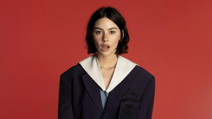 Portrait of a woman with short dark hair wearing a navy blazer with a white sailor-style collar, against a solid red background.