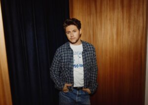 Young man leaning against a wooden wall wearing a blue plaid shirt over a white graphic tee and jeans, with hands in pockets.