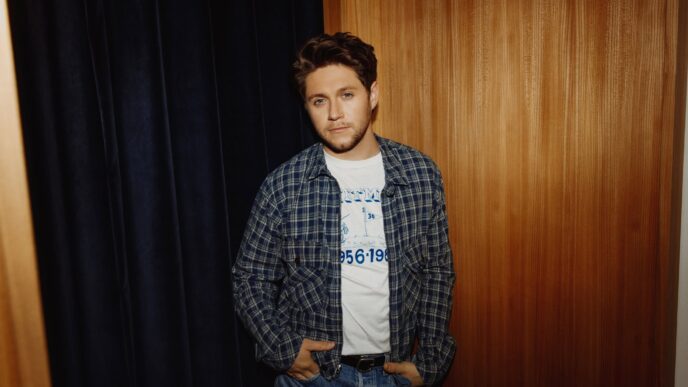Young man leaning against a wooden wall wearing a blue plaid shirt over a white graphic tee and jeans, with hands in pockets.