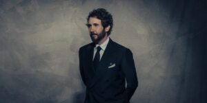 Man in a dark double-breasted suit and tie stands against a textured gray wall, looking to the left.