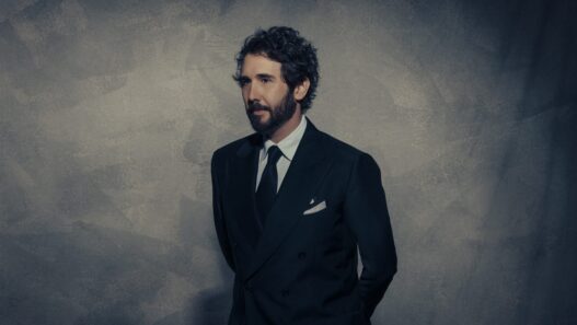 Man in a dark double-breasted suit and tie stands against a textured gray wall, looking to the left.