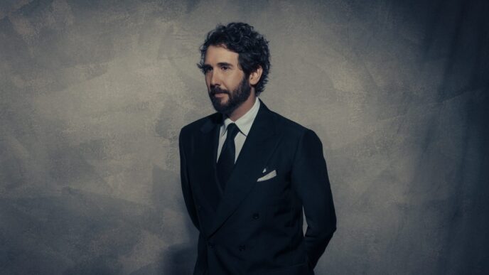 Man in a dark double-breasted suit and tie stands against a textured gray wall, looking to the left.