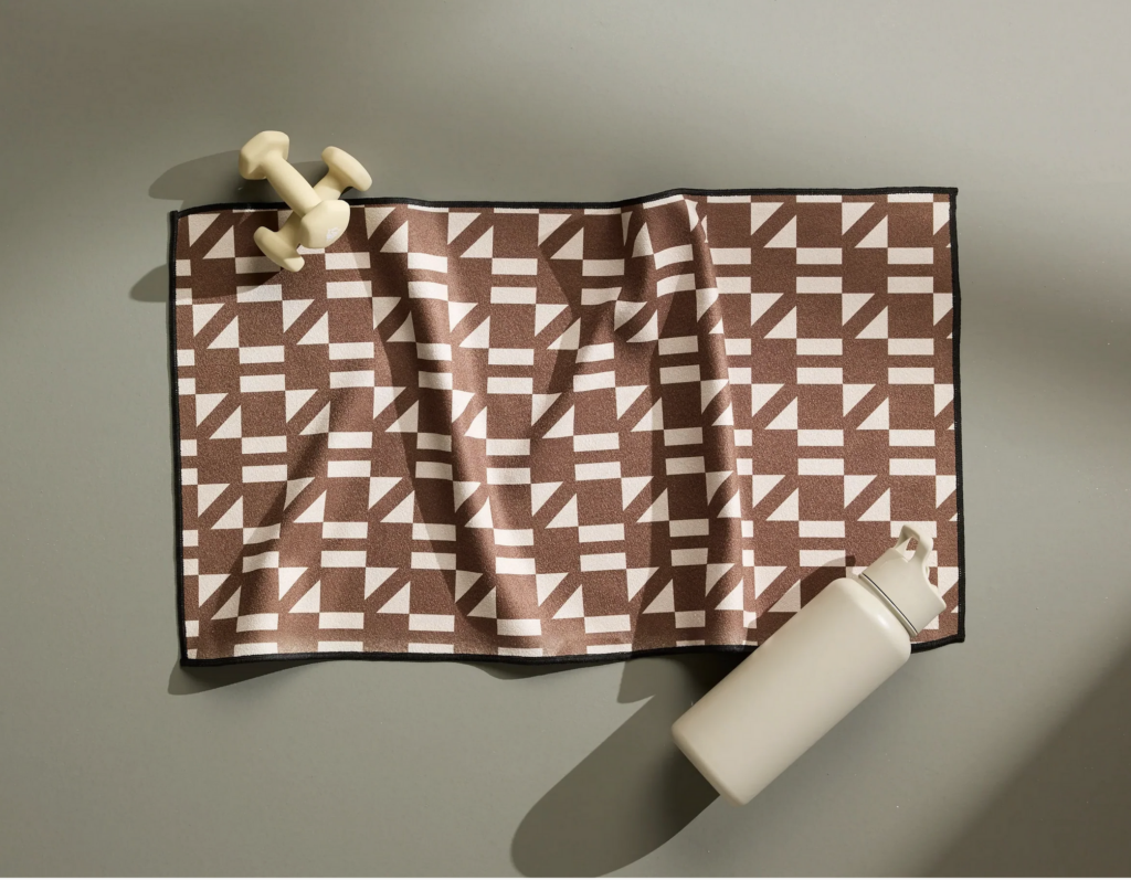 - Groovevolt Brown and white geometric patterned exercise mat on a gray floor, with a white water bottle nearby and light dumbbells at the edge.