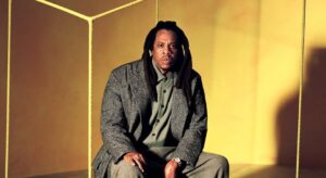 Man with dreadlocks in a grey suit and tweed overcoat seated on a chair inside a yellow studio, framed by rope edges around him.