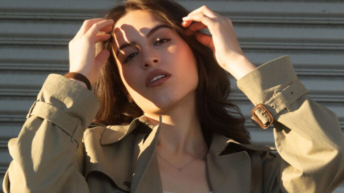 Young woman in a beige trench coat and white tank top, adjusting her hair in front of a corrugated metal backdrop.