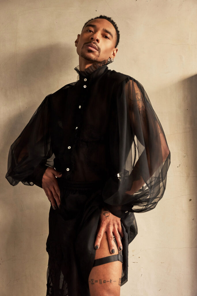 Person wearing a black sheer buttoned shirt with oversized mesh sleeves, posing against a beige wall.