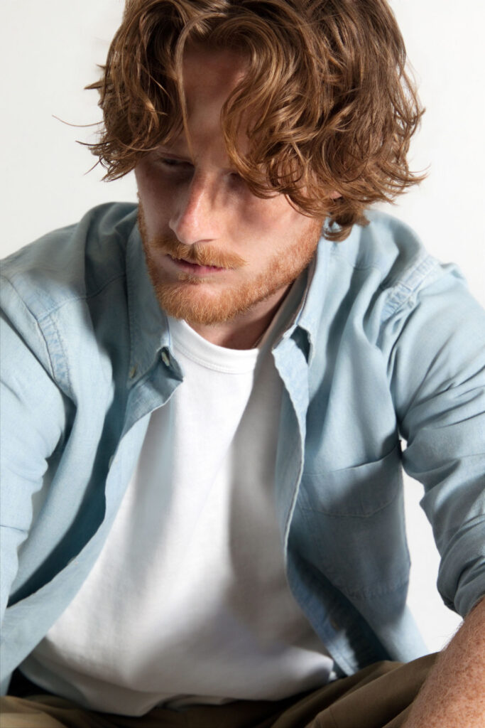 - Groovevolt Close-up portrait of a man with wavy red hair and a reddish beard, looking down while wearing a light blue shirt over a white T-shirt.