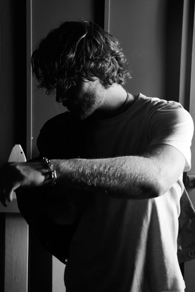 - Groovevolt Black-and-white portrait of a man with tousled hair, leaning against a wall with his arms crossed and head bowed downward.