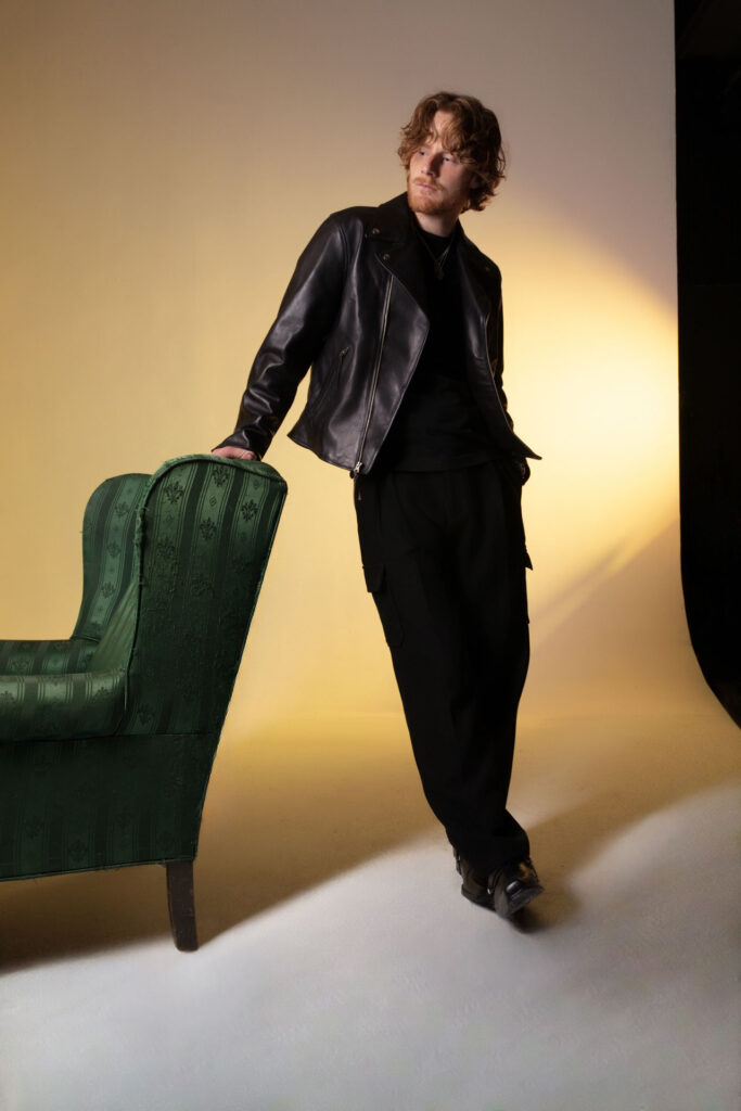 - Groovevolt Man with wavy red hair and beard in a black leather jacket leaning on a green patterned chair in a studio with warm yellow lighting.