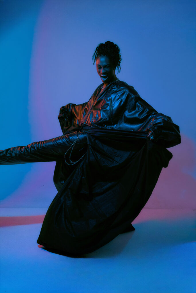 - Groovevolt Person in a shiny black coat dancing with arms spread, against a blue-pink gradient backdrop.