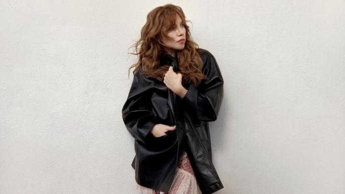 Woman with wavy red hair wearing an oversized black leather jacket over a pink dress, posing against a pale textured wall.
