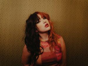 Portrait of a person with split black and red hair, bold makeup, wearing a colorful top, against a geometric patterned wall.