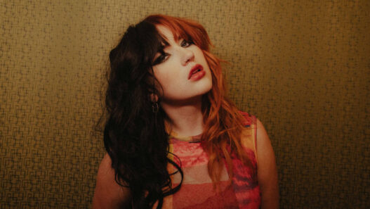 Portrait of a person with split black and red hair, bold makeup, wearing a colorful top, against a geometric patterned wall.