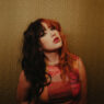 Portrait of a person with split black and red hair, bold makeup, wearing a colorful top, against a geometric patterned wall.