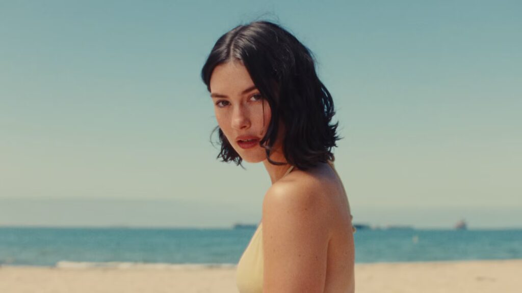 - Groovevolt Woman with short dark hair in a yellow bikini standing on a sandy beach with the sea in the background.