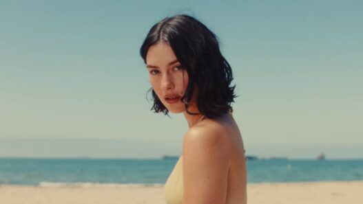Woman with short dark hair in a yellow bikini standing on a sandy beach with the sea in the background.