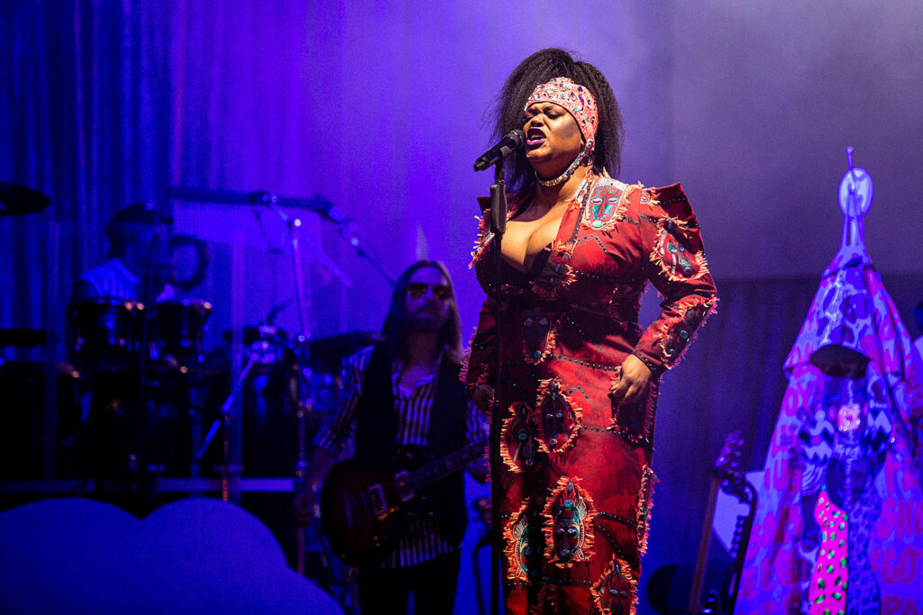 - Groovevolt Singer on stage wearing a vibrant red patterned outfit, singing into a microphone with blue stage lights and a backing band in the background.
