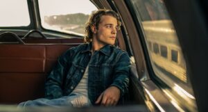 Young man in a denim jacket sits in the backseat of a vintage car, gazing out the window at sunset.