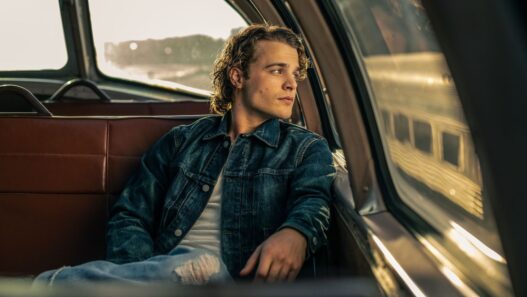 Young man in a denim jacket sits in the backseat of a vintage car, gazing out the window at sunset.