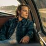 Young man in a denim jacket sits in the backseat of a vintage car, gazing out the window at sunset.
