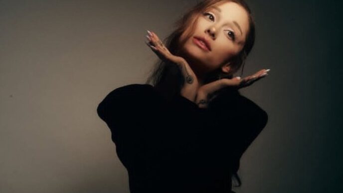 Portrait of a woman with long hair wearing a black top, resting her chin on her hands in a playful pose against a neutral studio background.