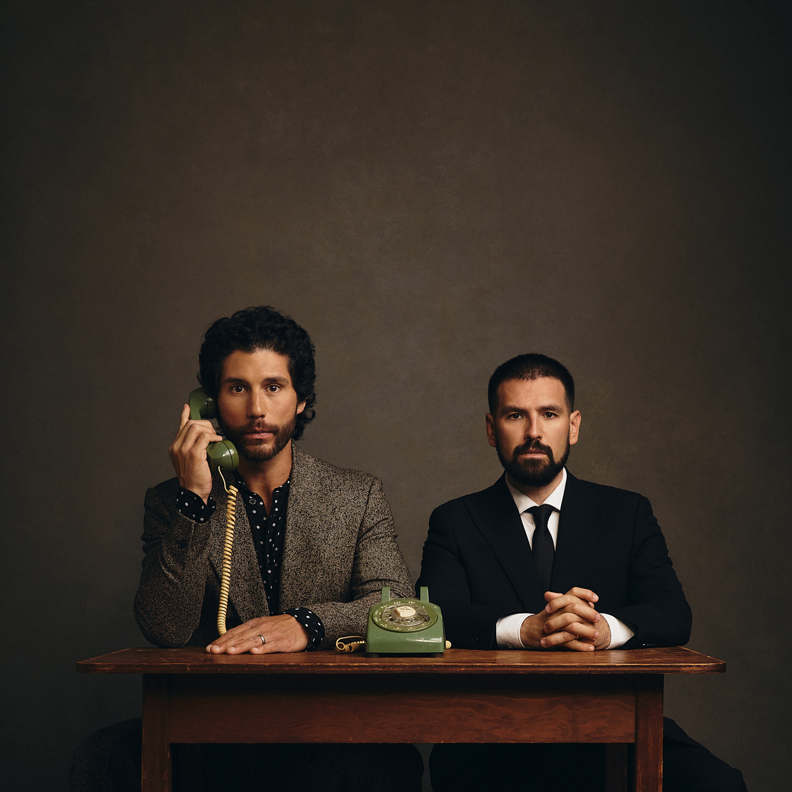 Two men sit at a wooden desk with vintage green rotary phones; the left man talks into the phone while the right man looks ahead in a dark studio setting.