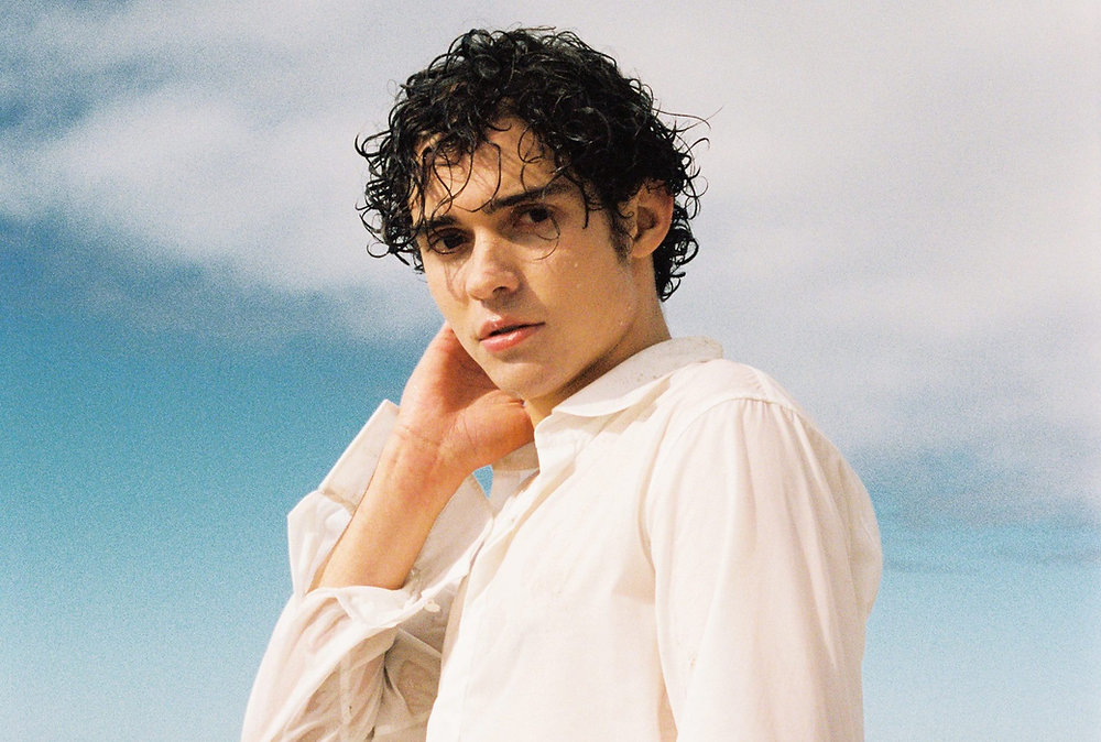 - Groovevolt Young person with curly dark hair in a white shirt, standing against a blue sky, hand near the neck.