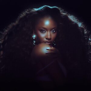 Glamour portrait of a woman with voluminous curly hair and blue backlighting, hand resting near her chin, looking at the camera.