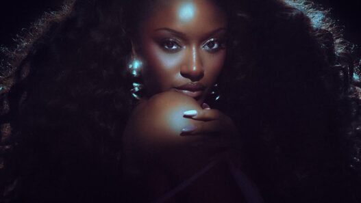 Glamour portrait of a woman with voluminous curly hair and blue backlighting, hand resting near her chin, looking at the camera.