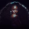 Glamour portrait of a woman with voluminous curly hair and blue backlighting, hand resting near her chin, looking at the camera.