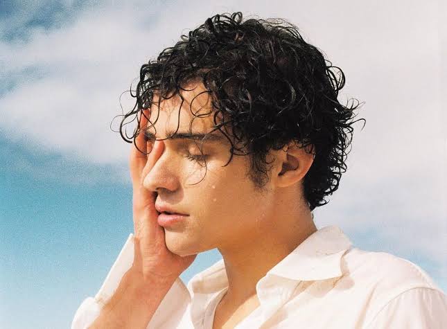 - Groovevolt Young man with curly, wet hair resting his cheek on his hand, wearing a white shirt outdoors under a blue sky.