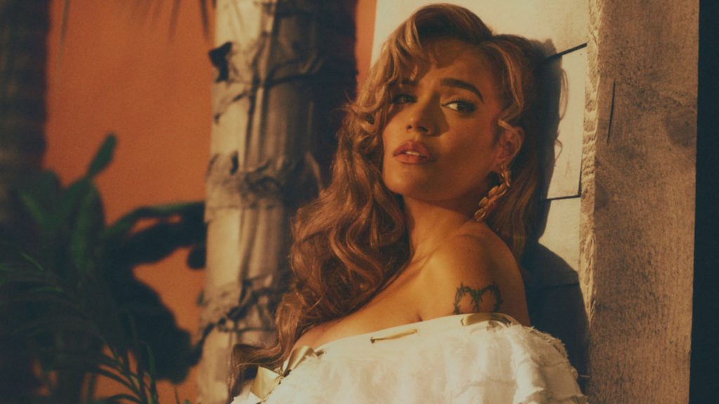 Woman with wavy hair leaning against a textured pillar in warm lighting, wearing a torn white off-shoulder dress; palm-tree tattoo on her chest.