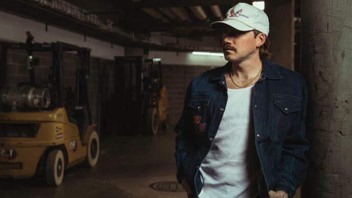 Man leaning against a concrete pillar in a dim industrial warehouse, wearing a light blue cap, denim jacket, and white shirt.