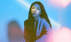 Portrait of a woman with long dark hair wearing a blazer, lit by blue and pink stage lighting.