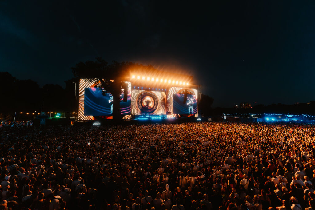 Massive outdoor concert crowd at night watching a brightly lit stage with circular LED visuals.