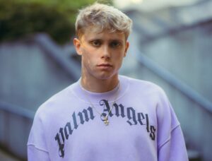 Portrait of a young man with short blond hair wearing a lavender sweatshirt with Gothic 'Los Angeles' text and a gold pendant necklace, outdoors.