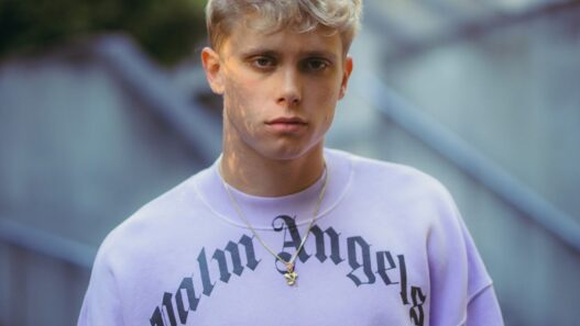 Portrait of a young man with short blond hair wearing a lavender sweatshirt with Gothic 'Los Angeles' text and a gold pendant necklace, outdoors.