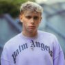 Portrait of a young man with short blond hair wearing a lavender sweatshirt with Gothic 'Los Angeles' text and a gold pendant necklace, outdoors.