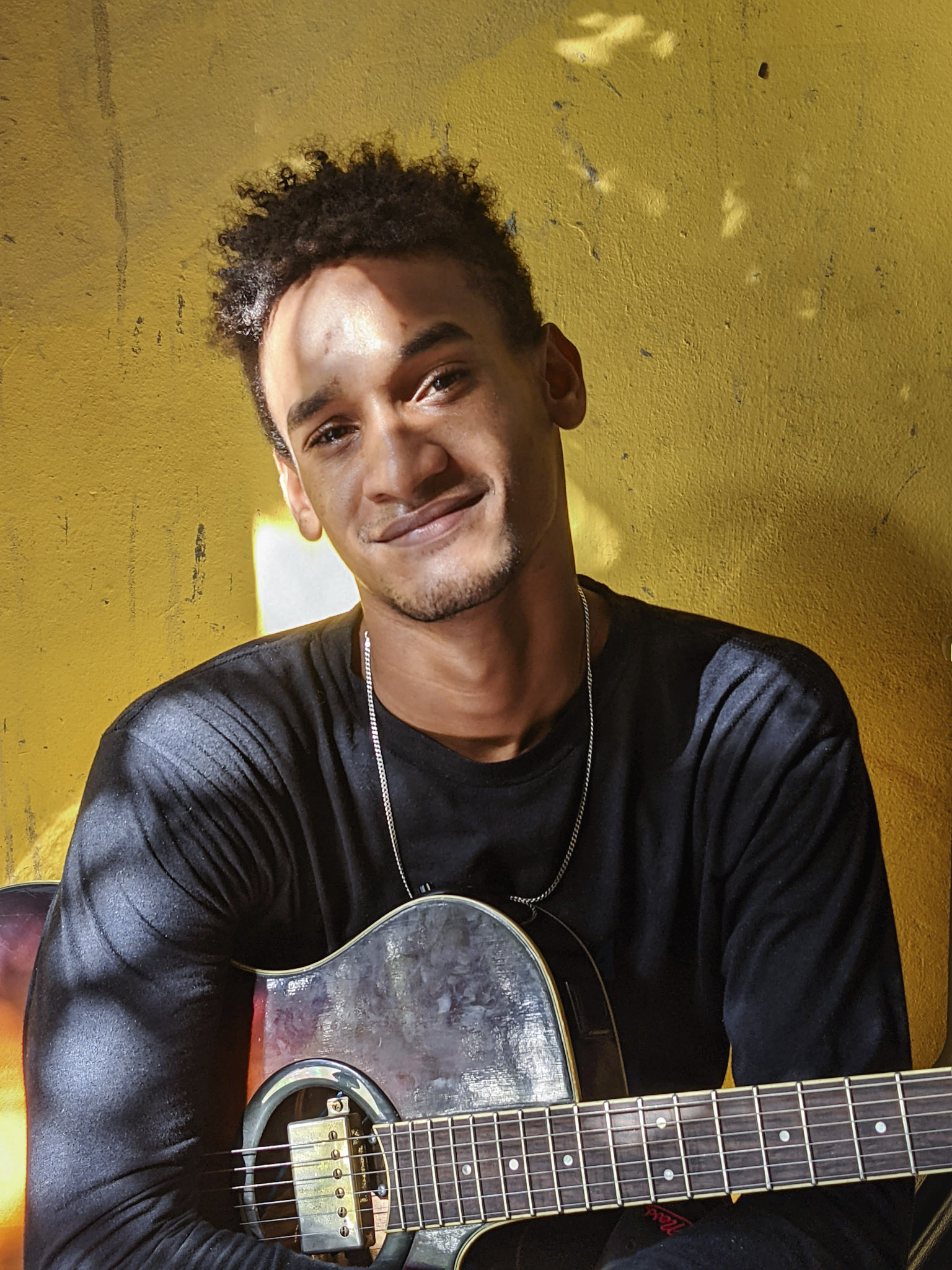 - Groovevolt Young man with short curly hair and a black shirt smiles while holding a guitar against a bright yellow wall.