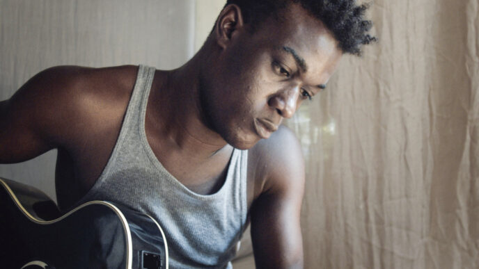 Young man in a gray tank top playing a dark acoustic guitar, looking down at the fretboard.