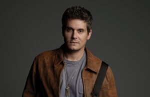 Portrait of a man with short dark hair wearing a brown leather jacket and gray tee, against a dark backdrop.
