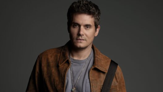 Portrait of a man with short dark hair wearing a brown leather jacket and gray tee, against a dark backdrop.