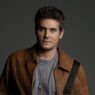 Portrait of a man with short dark hair wearing a brown leather jacket and gray tee, against a dark backdrop.