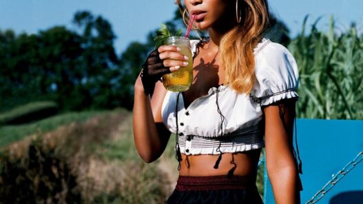 Young woman with blond highlights wearing a white cropped top and patterned shorts, sipping lemonade through a pink straw on a blue bench outdoors.