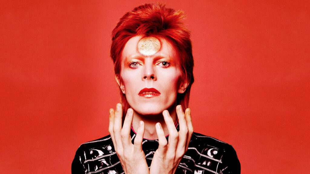 Portrait of a person with bright red hair and red lipstick against a red background, wearing a black studded jacket.