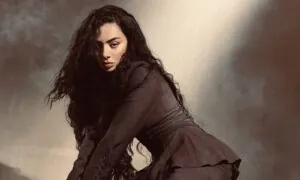 Woman with long dark curly hair in a brown coat, leaning forward against a muted studio backdrop.